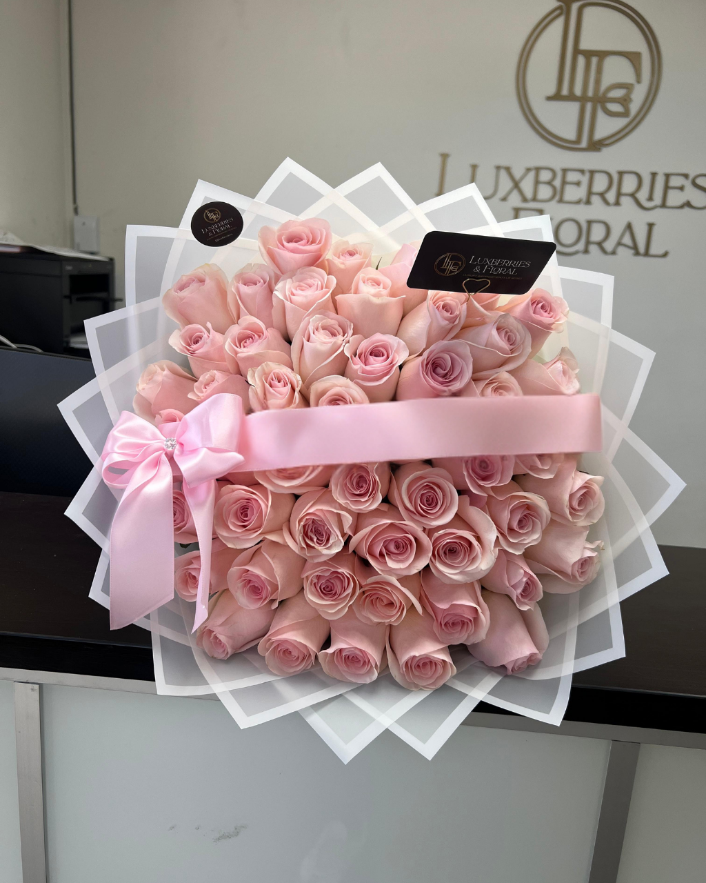 Bouquet of pink roses with a pink ribbon on a white stand, branded 'Luxberries Floral'.