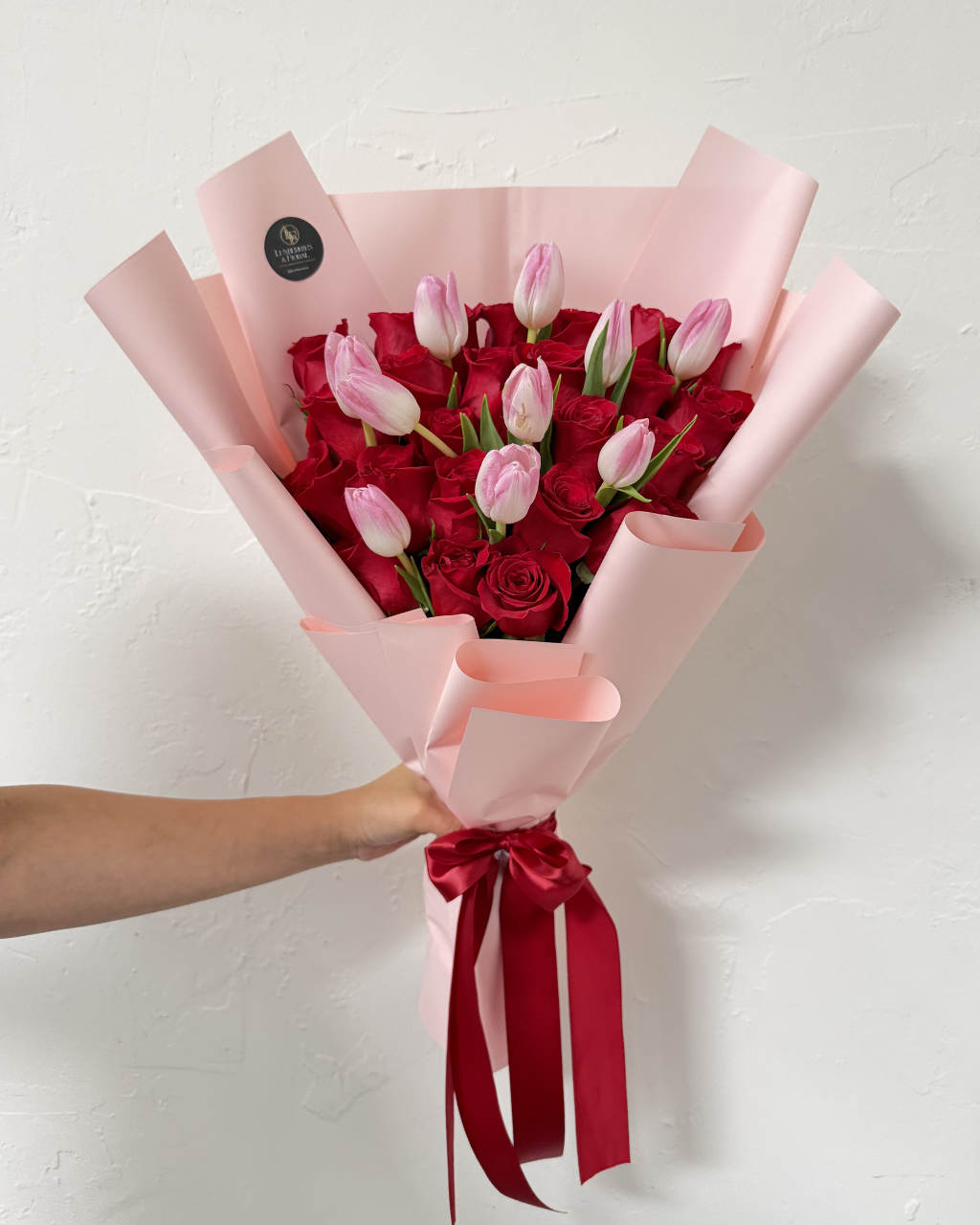 Bouquet of red and pink roses wrapped in pink paper with a red ribbon, held by a person against a white background.