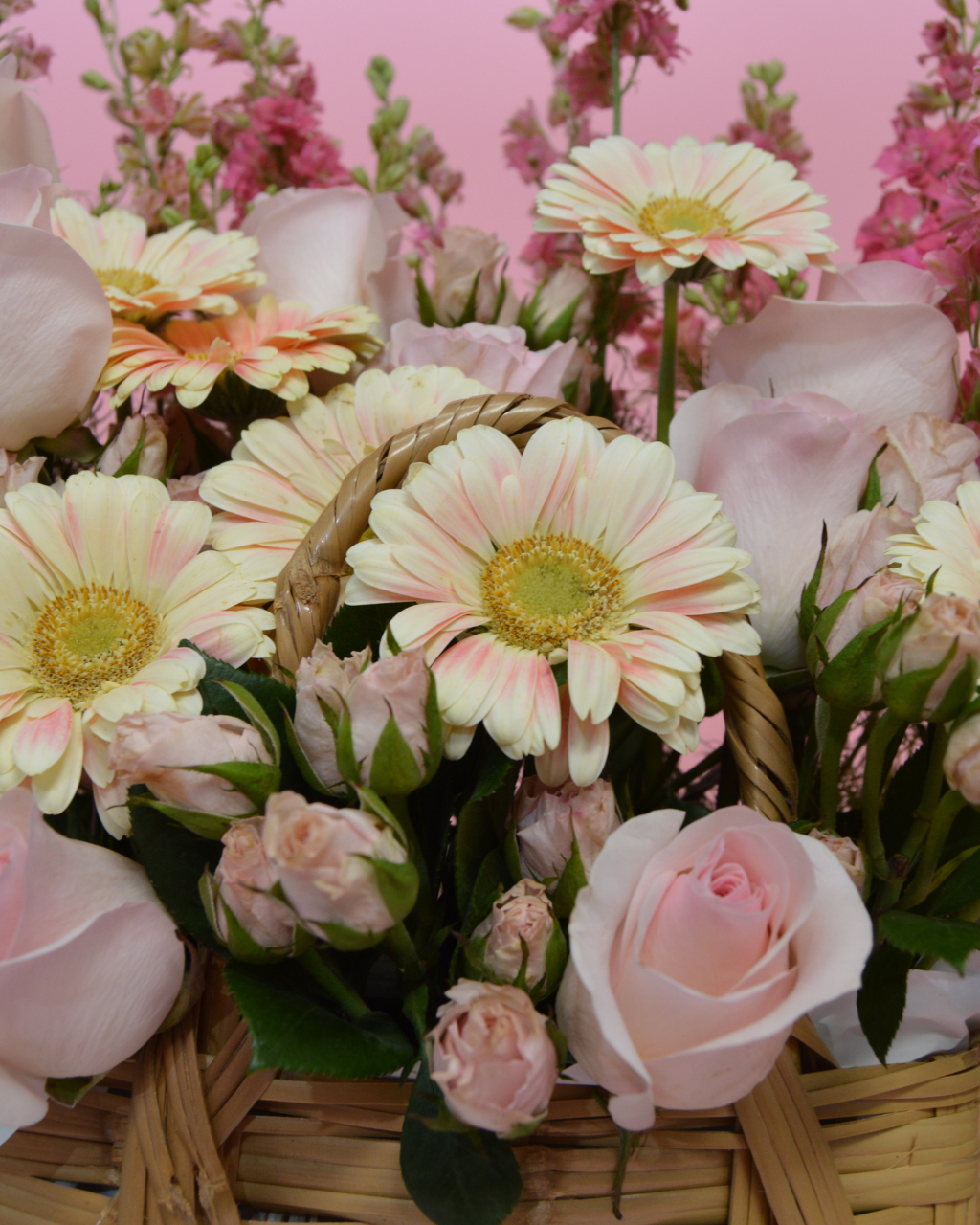 Bouquet of flowers including roses and daisies in a woven basket with a pink background