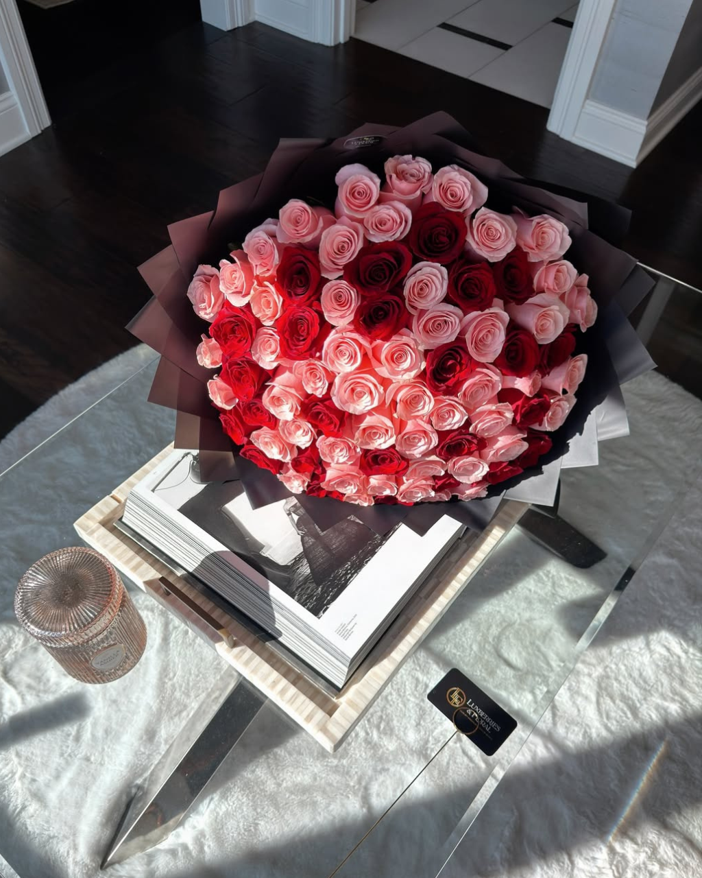 A large bouquet of pink and red roses wrapped in dark paper, placed on a glass table with books and a small card in a bright indoor setting.