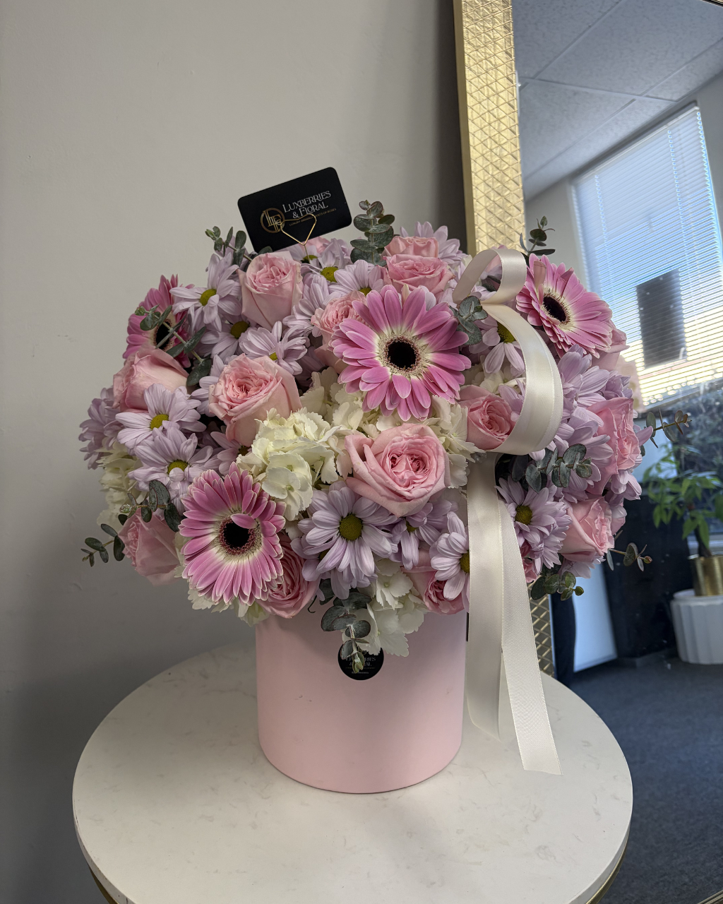 Bouquet of pink and white flowers in a pink box with a white ribbon on a white surface.