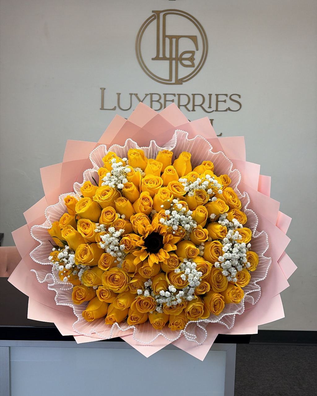 A large bouquet of yellow roses and a sunflower wrapped in pink paper, displayed in front of a wall with the LUVBERRIES logo.