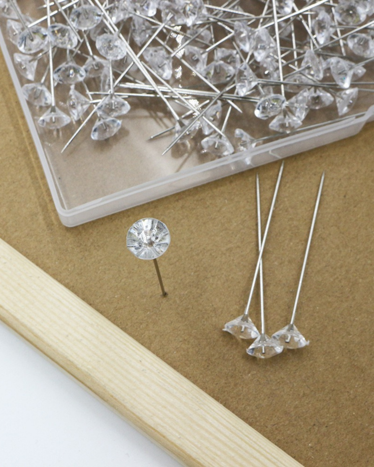 Clear faceted gemstone stud pins with long silver wires, shown in a plastic container and scattered on a brown surface next to a light wood block.