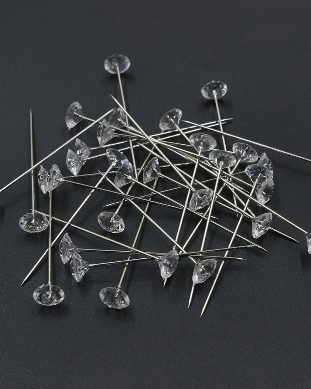 A pile of clear rhinestone push pins with long silver metal stems, scattered on a dark gray background.