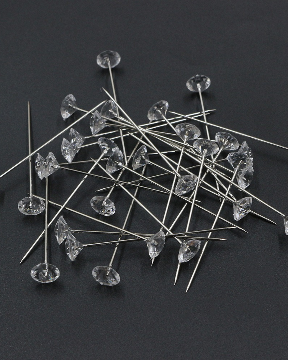 A pile of clear rhinestone push pins with long silver metal stems, scattered on a dark gray background.