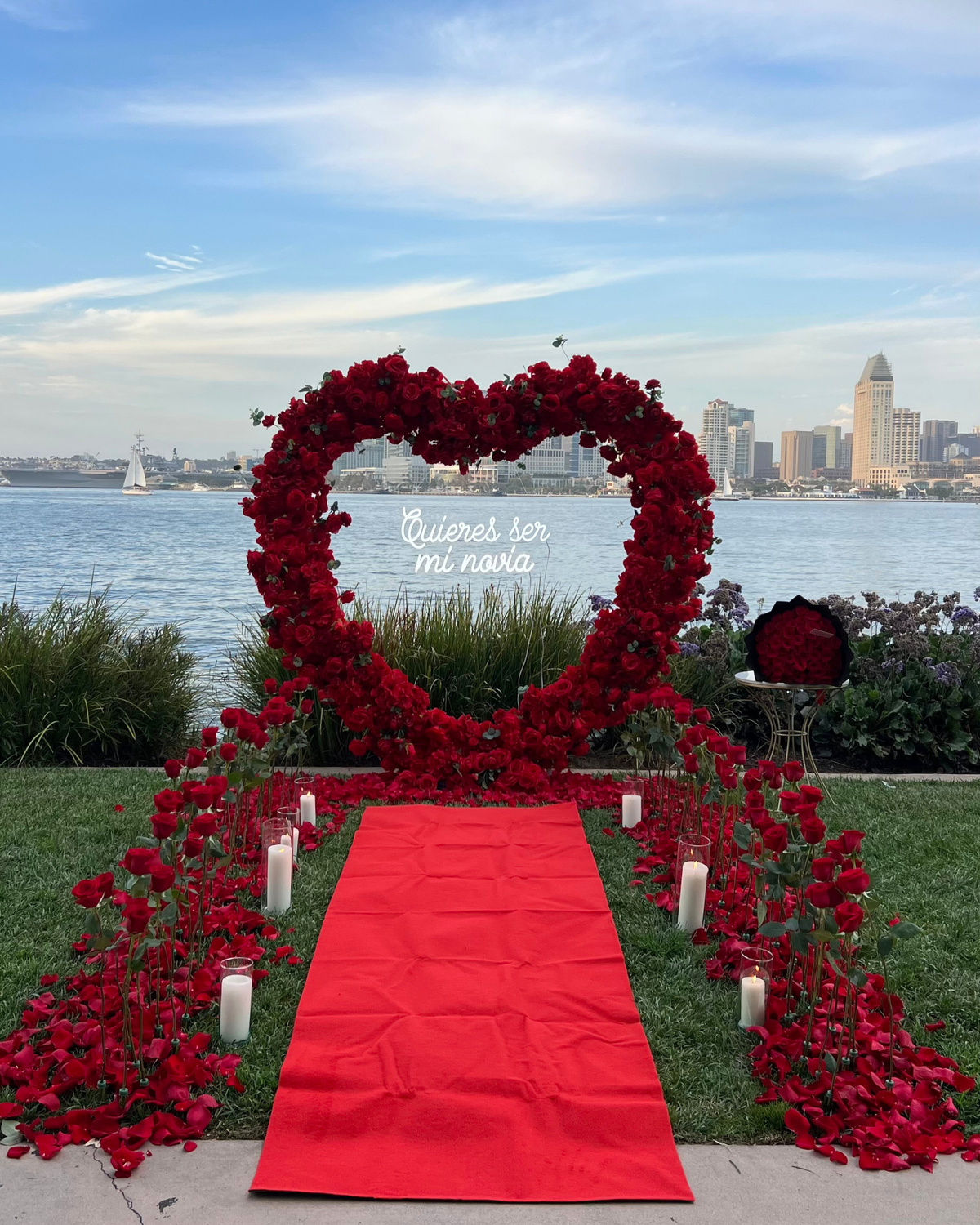 A romantic outdoor wedding or event setup by the water, featuring a large heart-shaped floral arch made of red roses with the Spanish text 'Quieres ser mi novia' on the arch, a bright red carpet leading down the center, red rose garlands lining the path, and white candles placed along the walkway. The background shows a city skyline across the water under a blue sky.