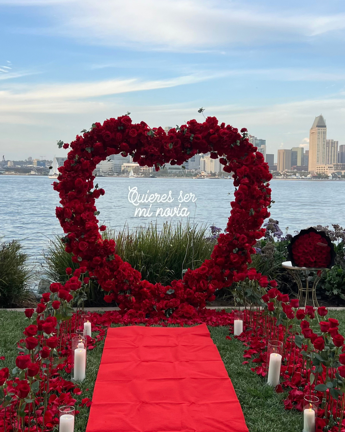 A romantic outdoor wedding or event setup by the water, featuring a large heart-shaped floral arch made of red roses with a white script sign reading 'Quienes ser mi novia.' A red carpet leads down the center, with red roses and white candles arranged along the grass.