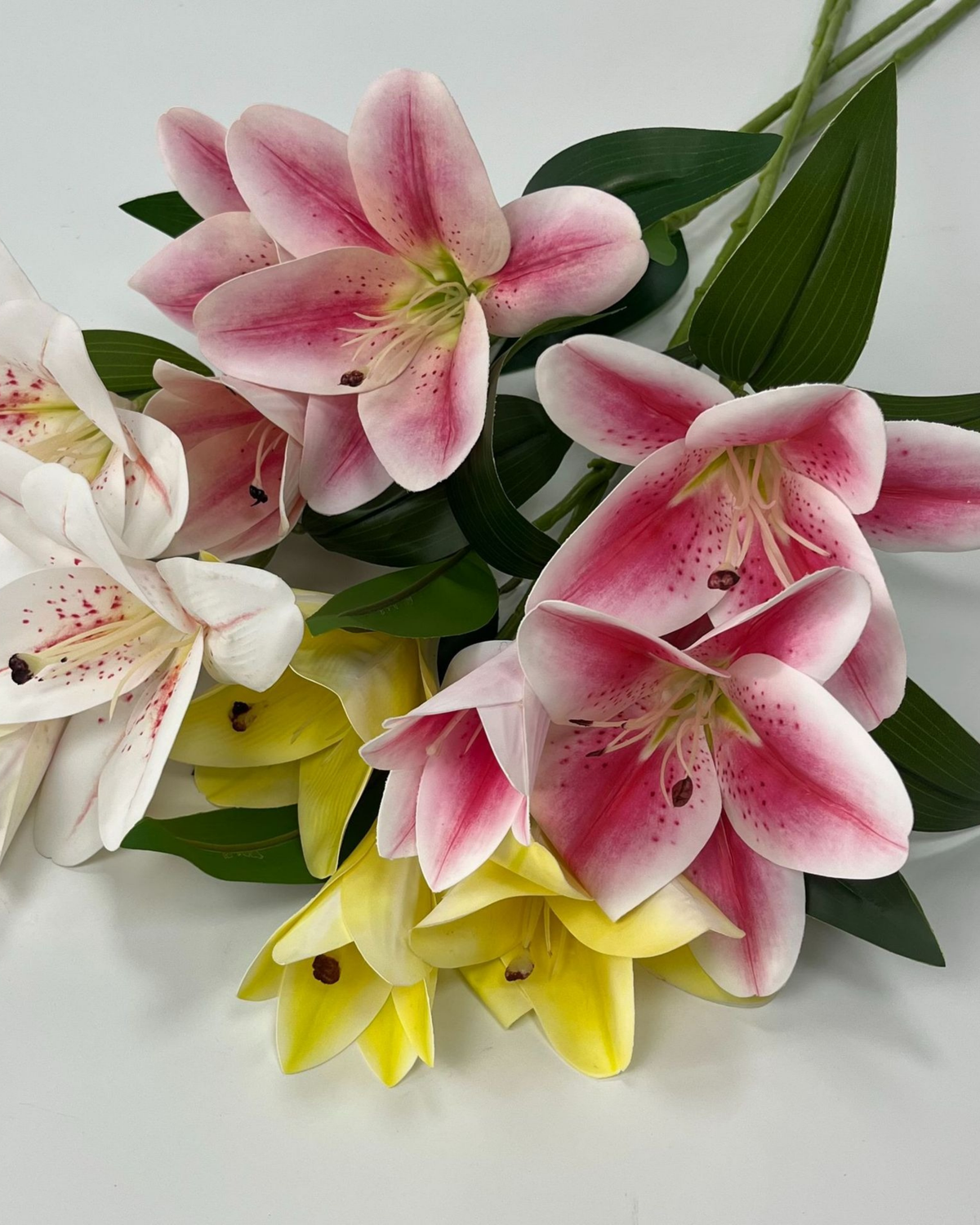 A bouquet of artificial lilies in pink, white, and yellow with green leaves, arranged on a plain white background.