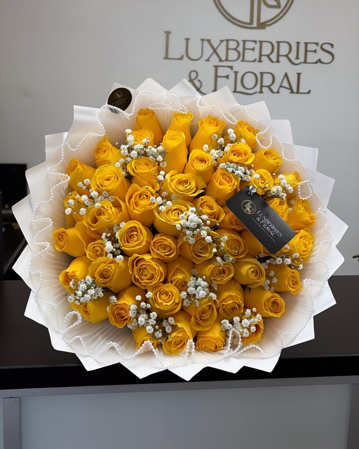A large bouquet of yellow roses wrapped in white paper, decorated with small white baby's breath flowers and a black Luxberries & Floral gift tag, displayed on a counter with the brand logo on the wall behind.