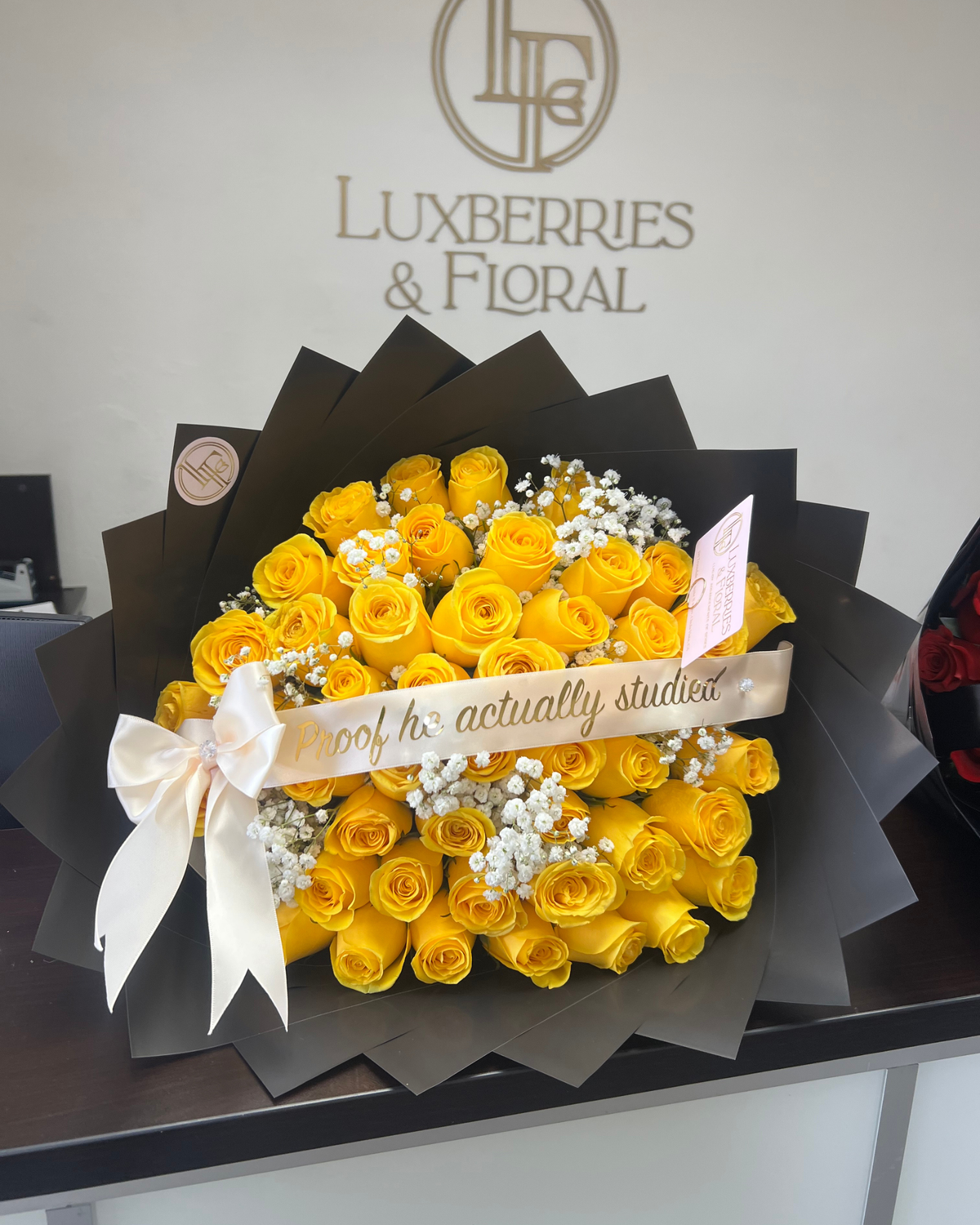 A large bouquet of yellow roses wrapped in black paper with a white ribbon and a gold banner that reads 'proof to actually studied'. The bouquet is displayed on a dark table against a white wall with the Luxberries & Floral logo.