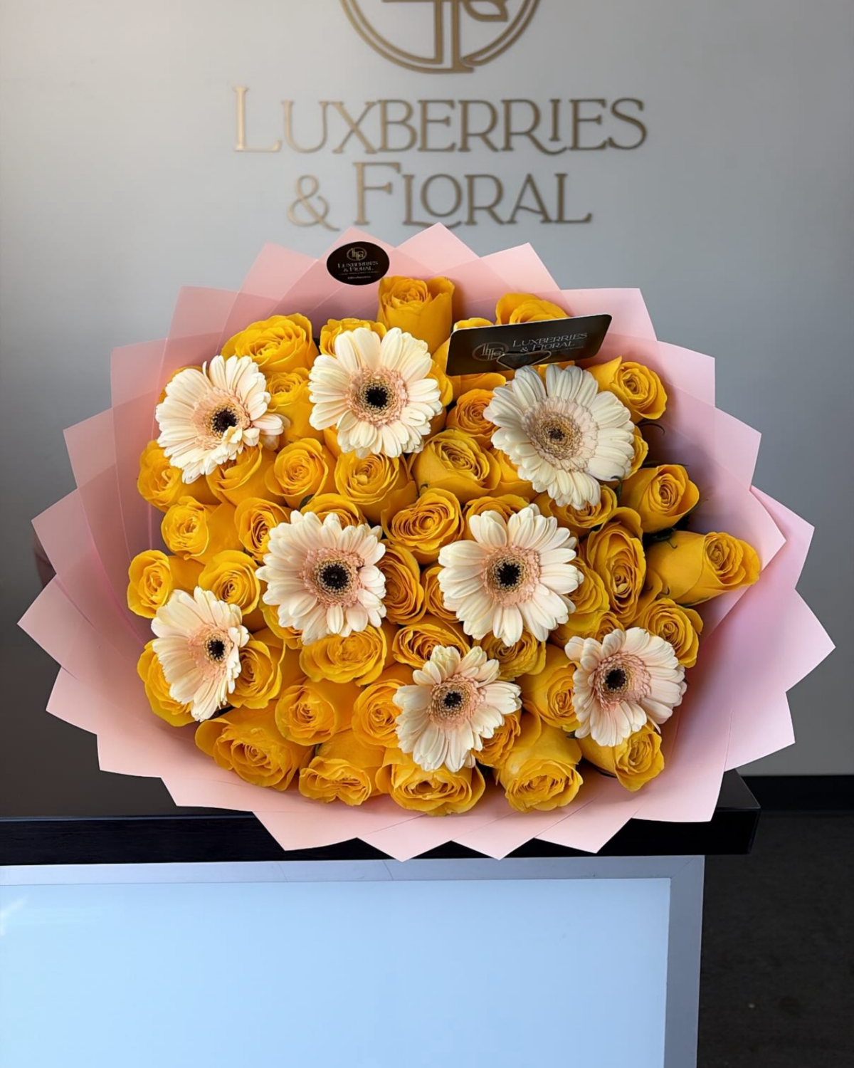 A bouquet of yellow roses and white/cream Gerbera daisy flowers wrapped in pink paper, displayed on a counter with the Luxberries & Floral logo on the wall behind it.