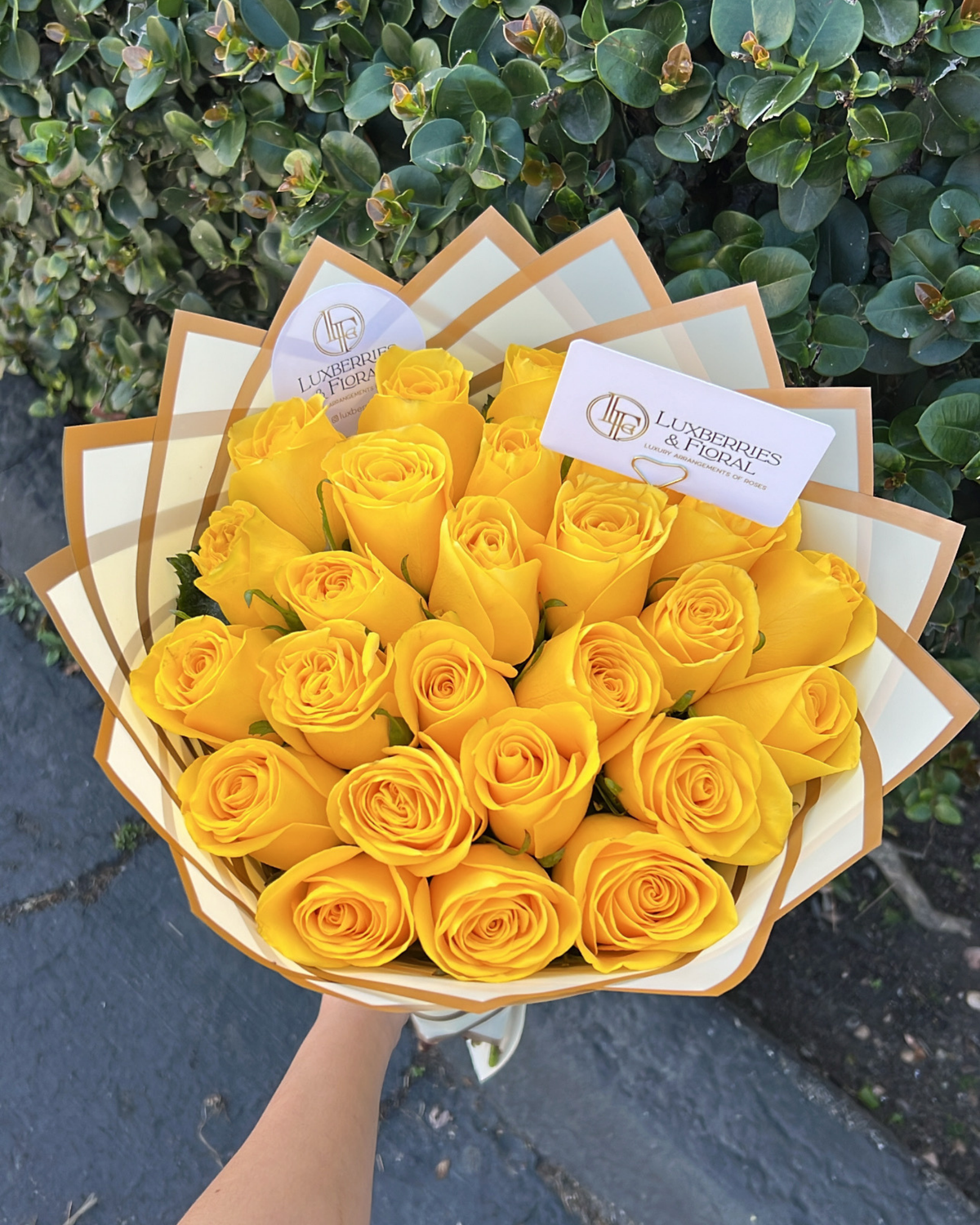 A bouquet of yellow roses wrapped in cream and gold paper, held by a hand against a background of green bushes and gray pavement. Two cards are visible, one showing the logo and name 'Londres & Froid'.