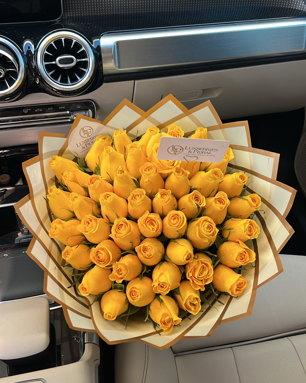 A large bouquet of yellow roses wrapped in cream paper with gold borders, placed on the center console of a car interior. Two small cards are visible at the top of the bouquet.