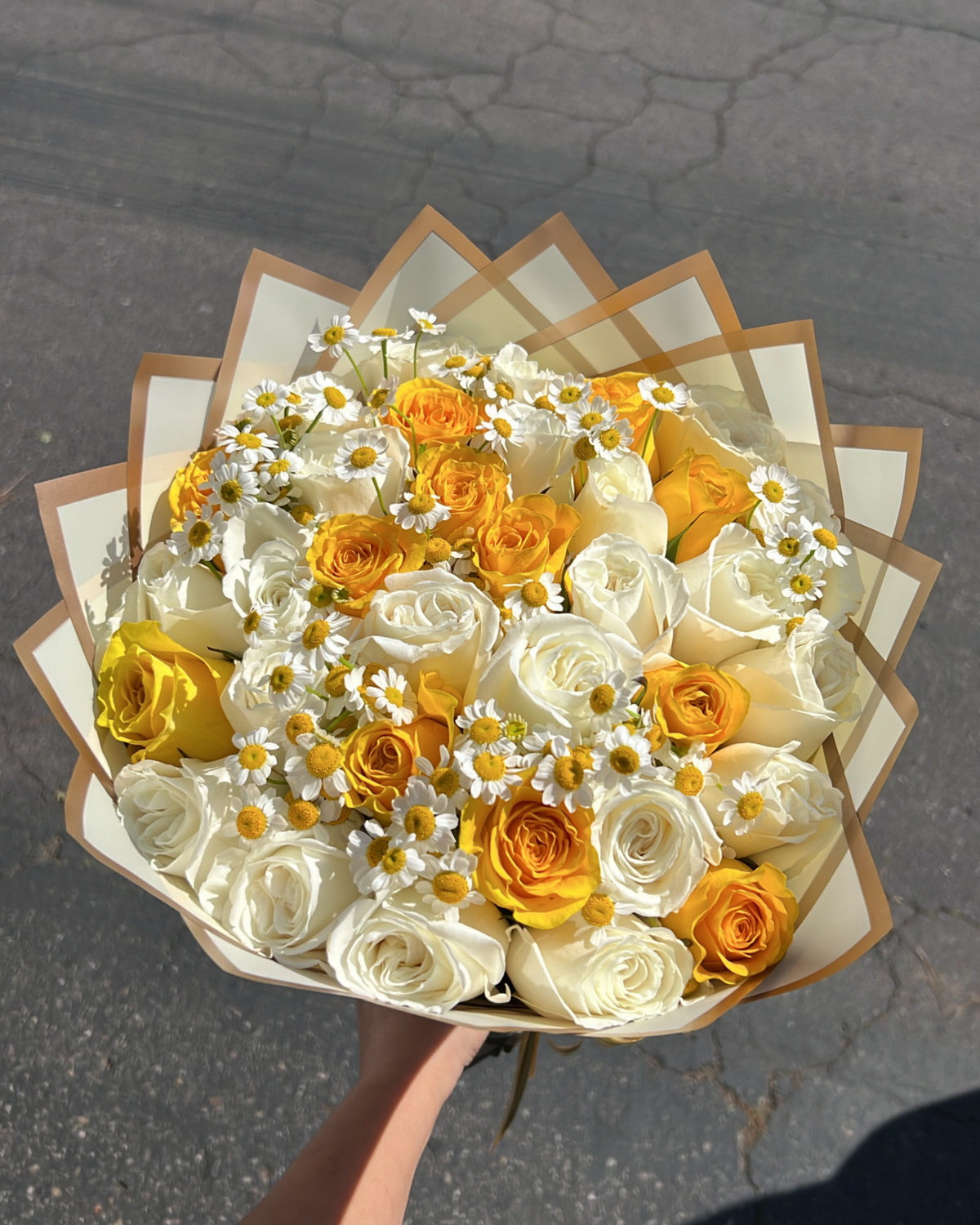 A bouquet of white and yellow roses with small white daisies, held in a hand against an outdoor asphalt background.