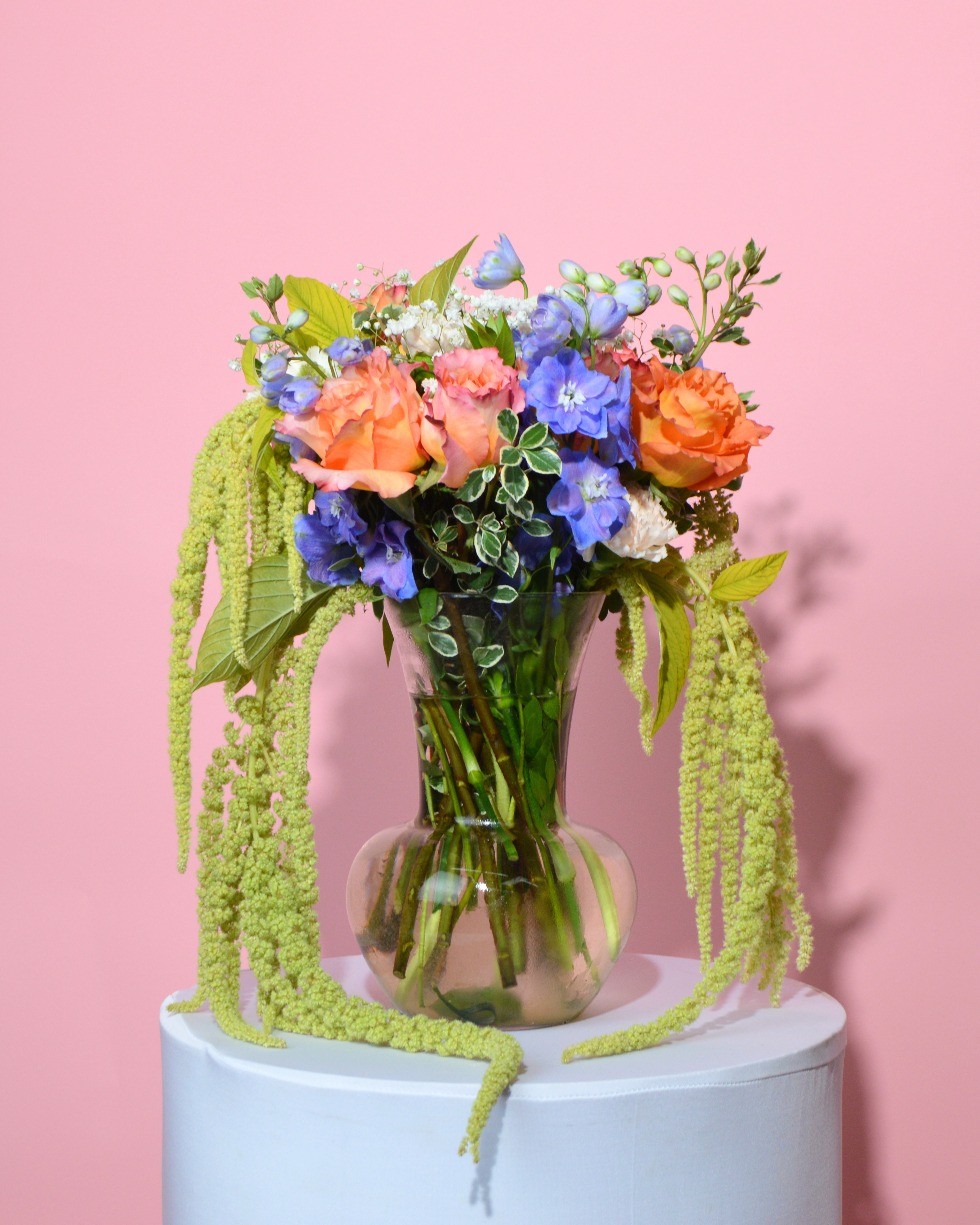 A vibrant artificial flower bouquet in a clear glass vase, featuring orange roses, blue delphiniums, white filler flowers, and green trailing agapanthus, displayed on a white pedestal against a solid pink background.