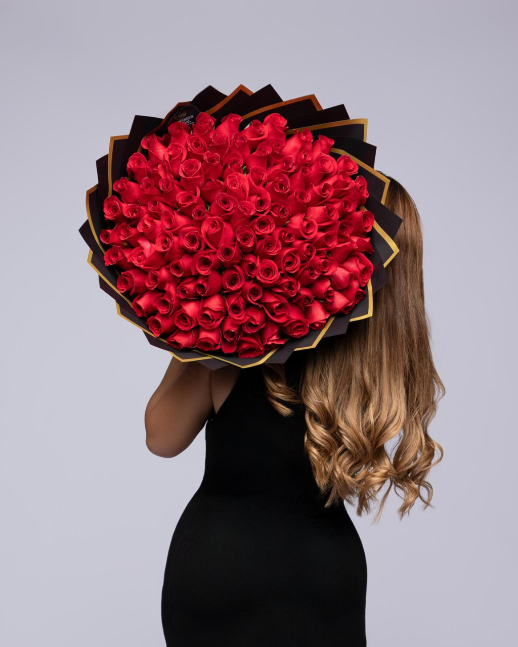 A woman in a black dress holds a large, round bouquet of red roses with black and gold wrapper behind her head, photographed against a solid light gray background.