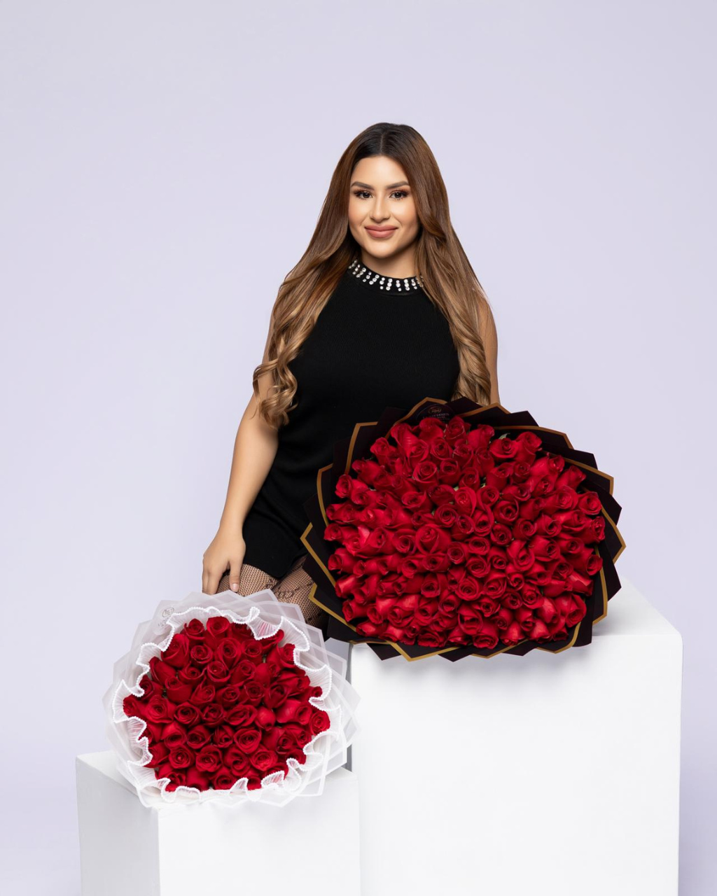 A woman in a black dress stands behind two large round arrangements of deep red roses on white pedestals against a light lavender studio background.