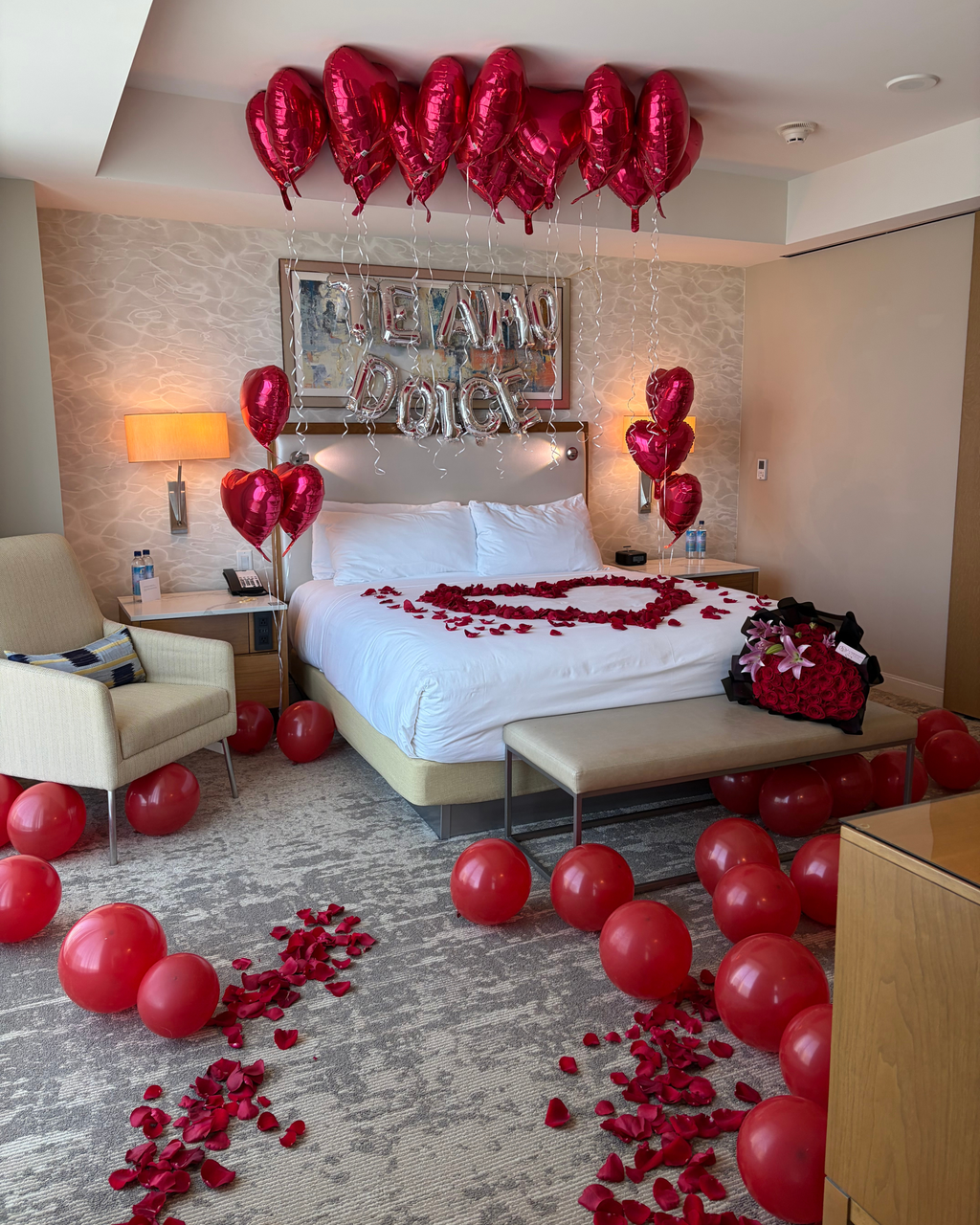 A romantic Valentine's Day themed hotel room decorated with red heart-shaped balloons, scattered red rose petals, and a heart-shaped balloon arrangement on the bed. A gift bag filled with red roses sits on the floor next to the bed.