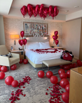 A romantic Valentine's Day themed hotel room decorated with red heart-shaped balloons, scattered red rose petals, and a heart-shaped balloon arrangement on the bed. A gift bag filled with red roses sits on the floor next to the bed.