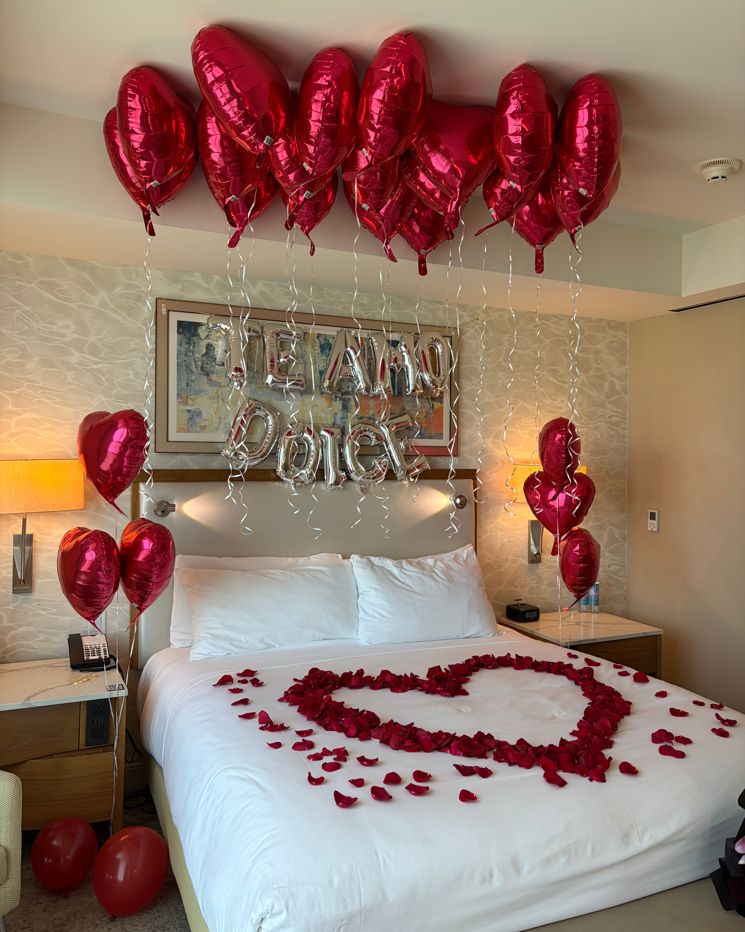 A romantic bedroom setup with a bed made of white linens, red rose petals arranged in a heart shape, metallic red heart balloons floating above, and silver balloon letters spelling names on the wall.