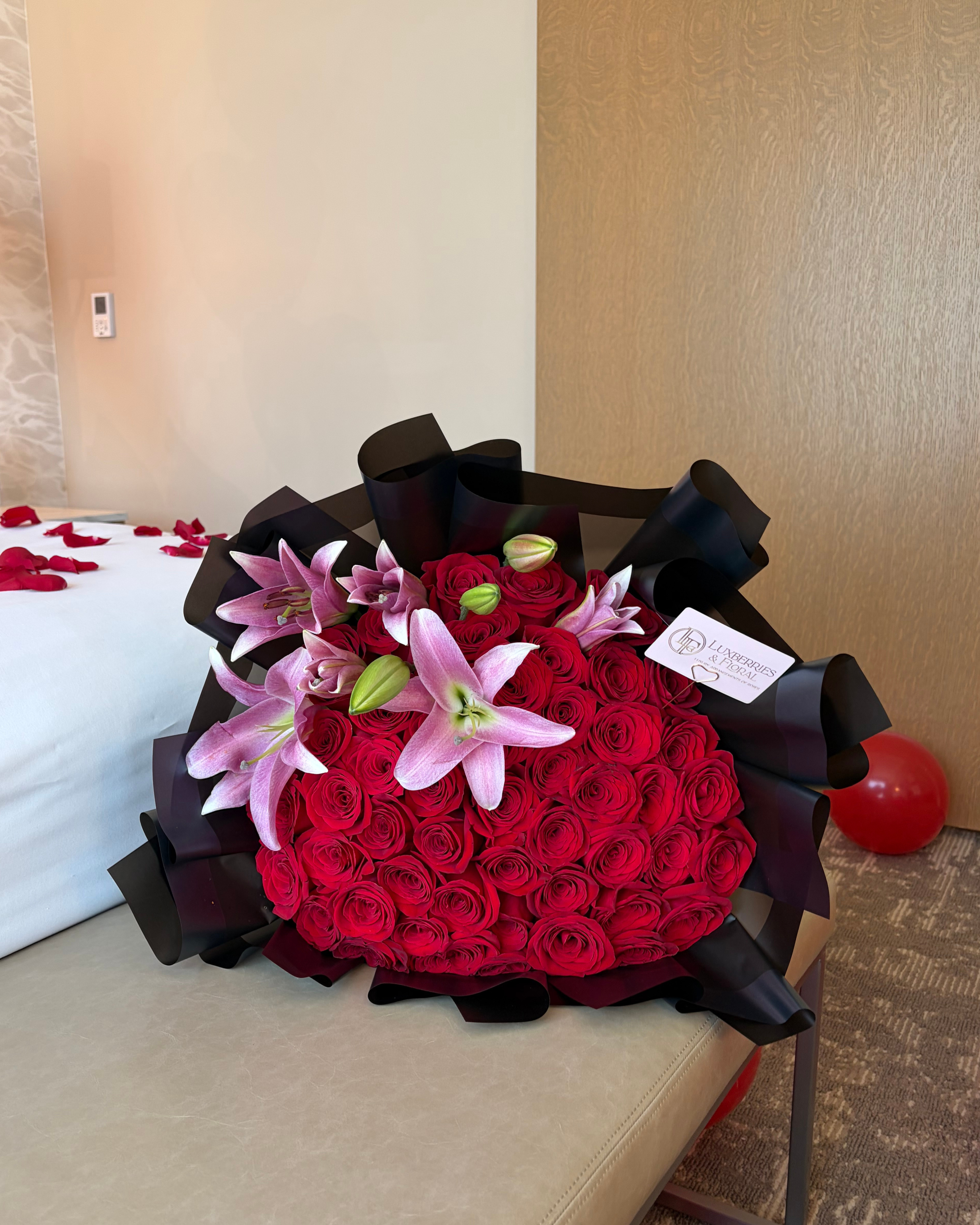 A large bouquet of red roses wrapped in black paper with pink lilies, placed on a beige bench in a room with a bed and wood-paneled wall in the background.