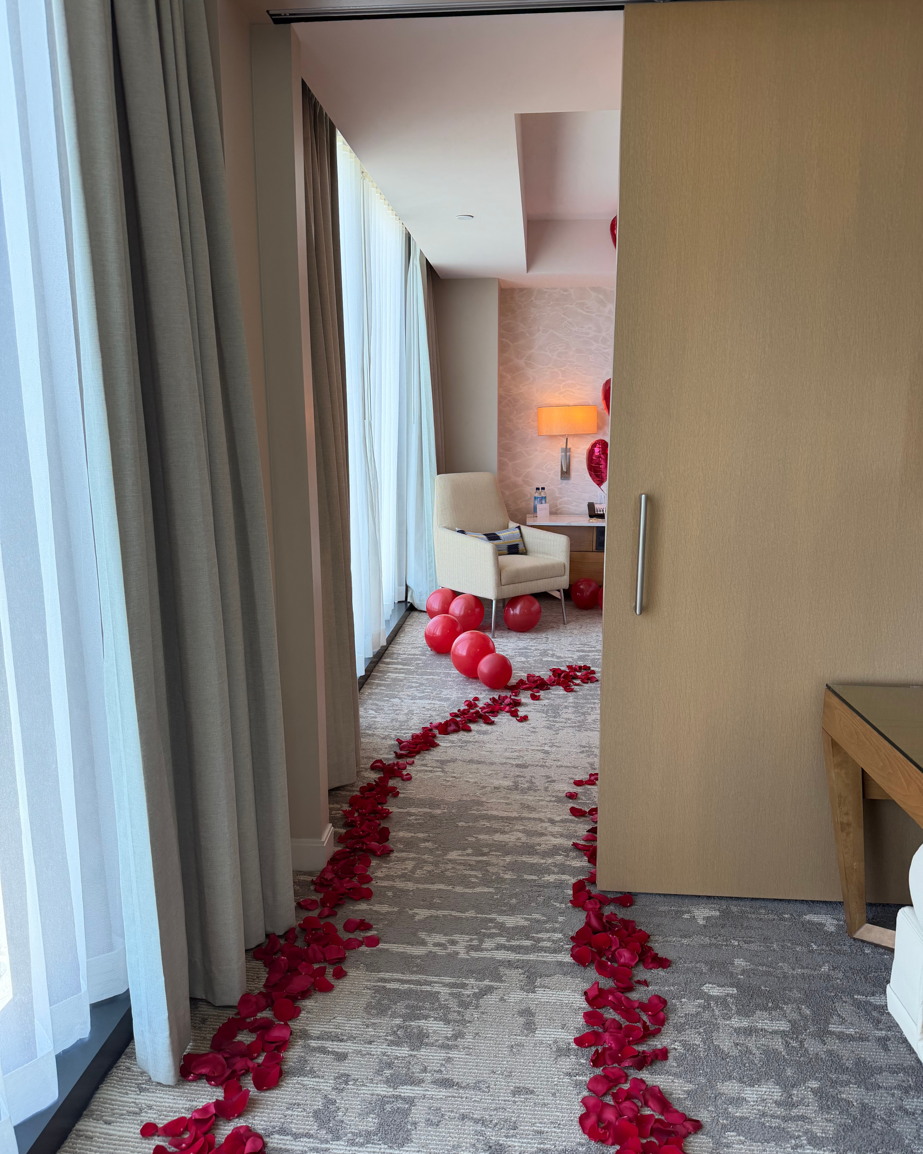 A romantic hotel room setup with a carpeted path lined with red rose petals leading from the foreground to a seating area decorated with red balloons. A sliding wooden door is open on the right, and curtains frame the left side of the scene.