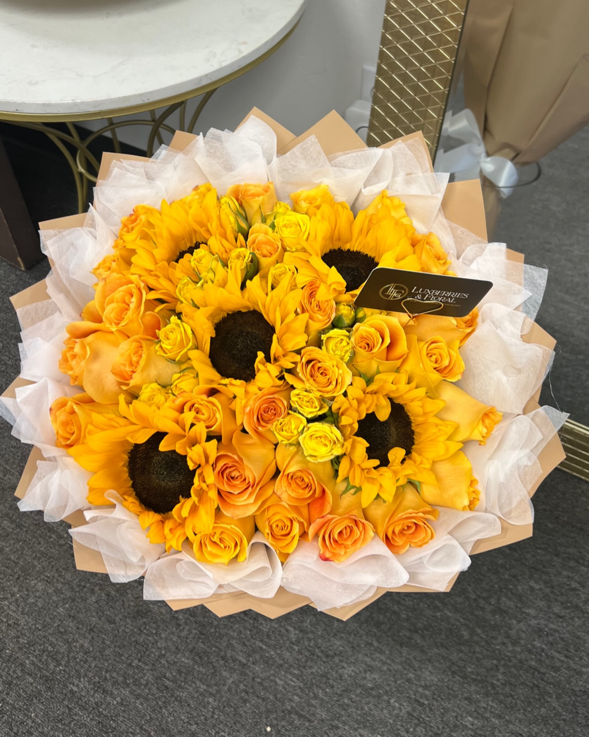 A bouquet of sunflowers and orange roses wrapped in white tissue paper and brown paper, with a small black card tag, photographed on a gray floor.