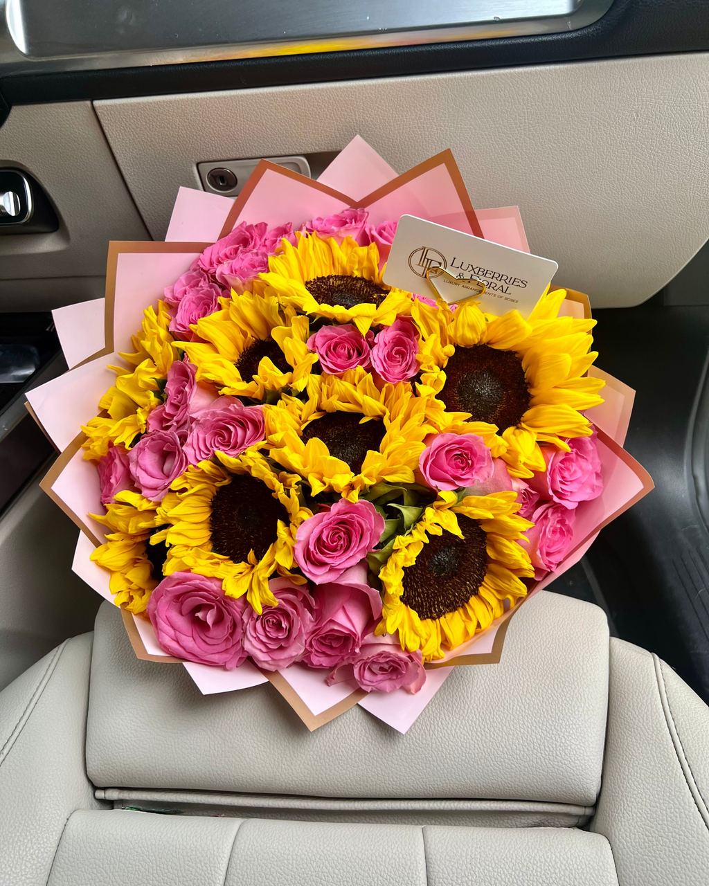 A large bouquet of yellow sunflowers and pink roses wrapped in pink paper, placed on the center console of a car with beige leather seats. A card reading 'Floristexpress Global' is visible.
