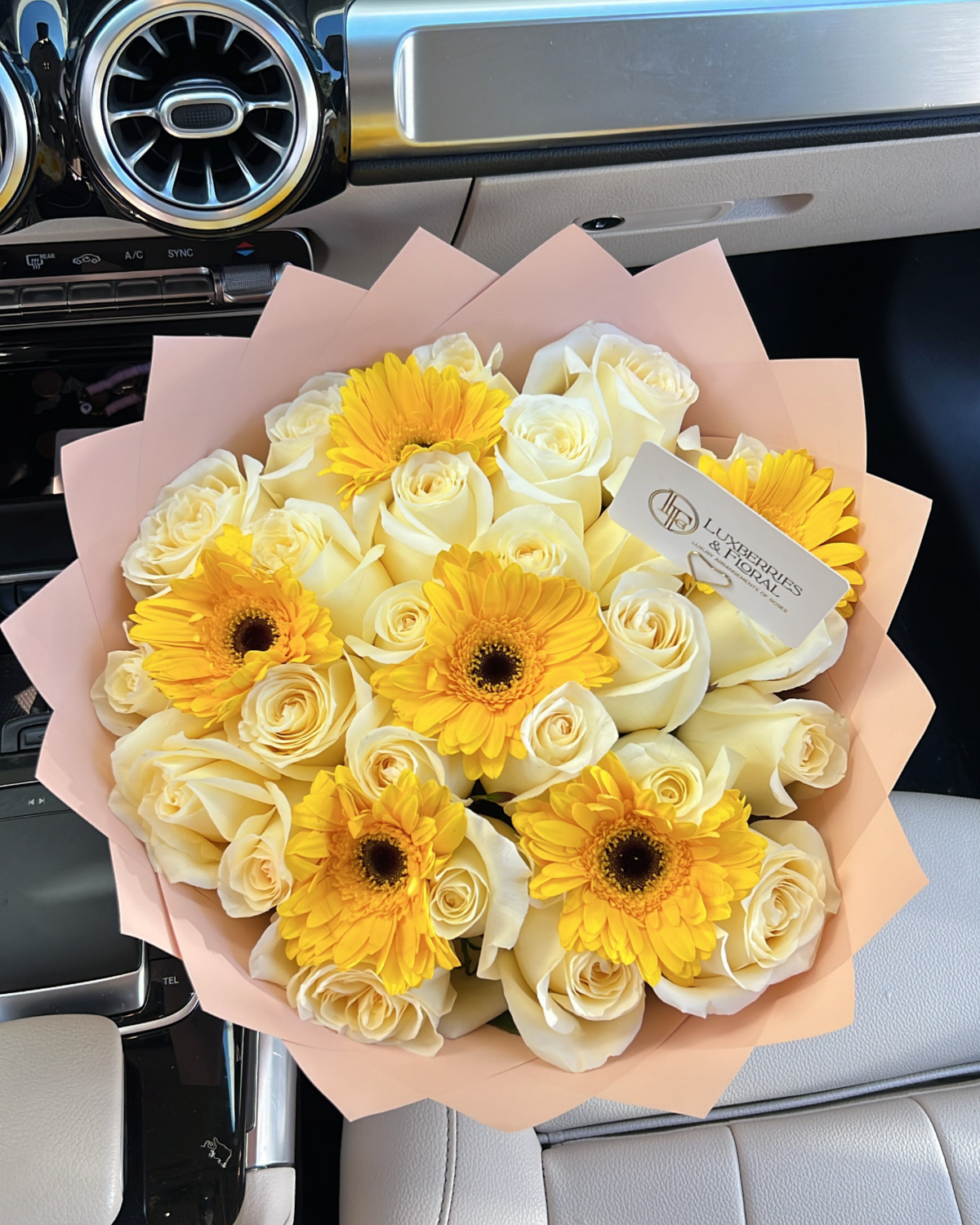 A bouquet of white and yellow flowers, including white roses and yellow gerbera daisies, wrapped in a light pink paper sleeve and placed inside a car interior.