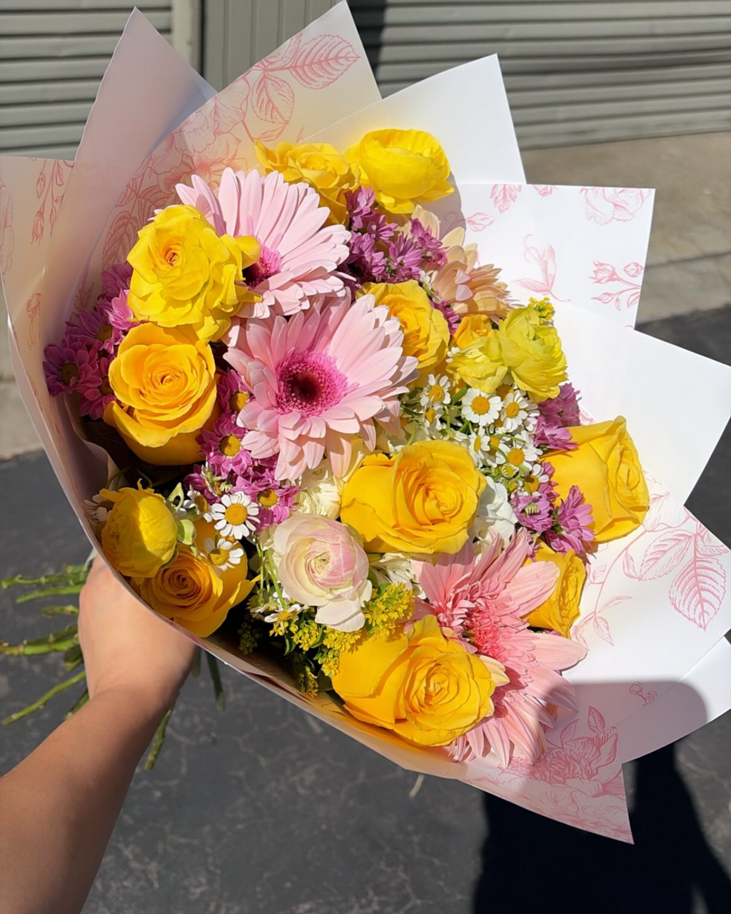 A hand holding a vibrant flower bouquet wrapped in white patterned paper. The bouquet features yellow roses, pink and yellow gerbera daisies, pink carnations, and small white daisies.