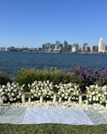 A wedding-style floral arrangement of white roses and white candles on a green lawn, with a body of water and a city skyline in the background under a clear blue sky.