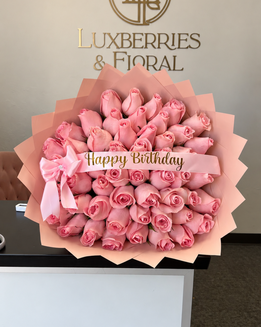 A large bouquet of pink roses wrapped in peach paper, tied with a pink ribbon that says 'Happy Birthday' in gold lettering. The bouquet is displayed on a counter in front of a wall with the brand name 'LUXBERRIES & FIORAL' in gold.