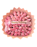 A round bouquet of pink roses wrapped in peach paper, tied with a pink ribbon bow and a white banner that reads 'Happy Birthday' in gold lettering, shown on a plain white background.